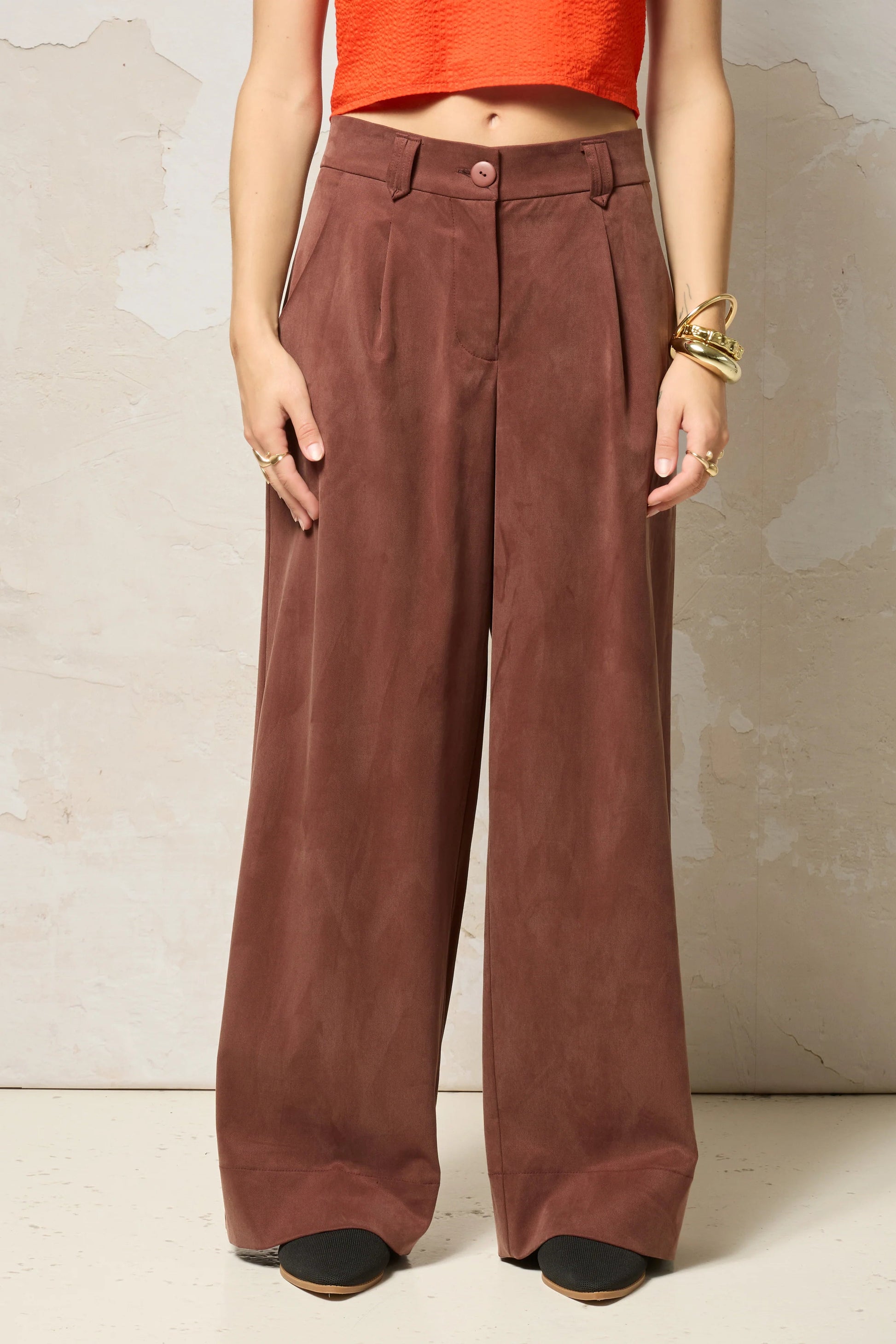 Waist down view of a woman wearing  the Chain pants from Cokluch in Cappuccino, with a slightly low waist, pointed belt loops, front pleats, and wide legs. She is wearing them with an orange top  and standing against a grey background. 