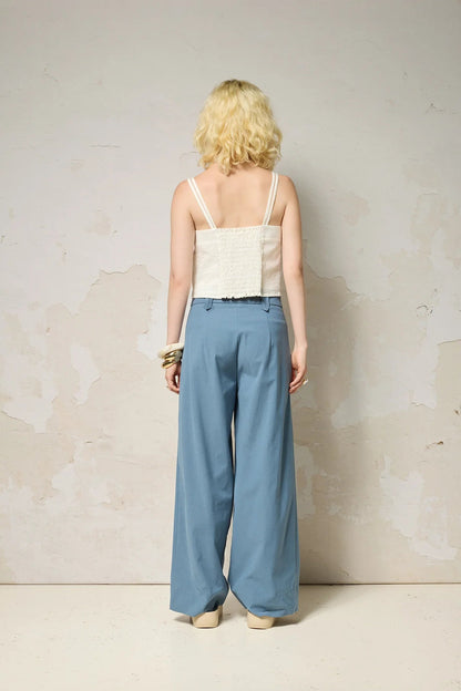 Back view of a woman wearing  the Chain pants from Cokluch in Ocean, with a slightly low waist, pointed belt loops, front pleats, and wide legs. She is wearing them with a white top  and standing against a grey background.