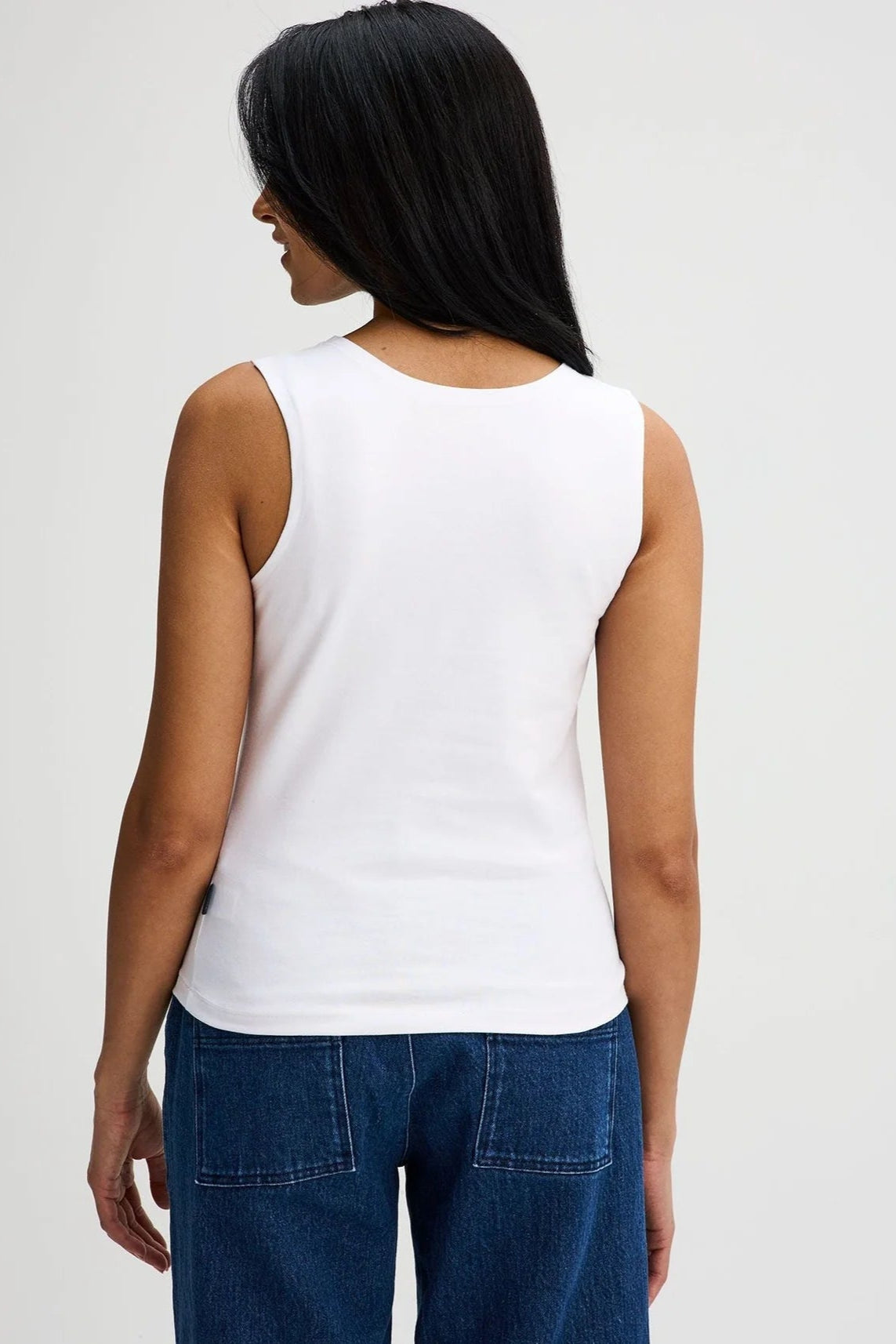 Back view of a woman wearing the Brompton Tank by Message Factory in White, a crew neck tank with wide straps and a slim fit in organic cotton. She is wearing it with jeans and standing in front of a white background. 