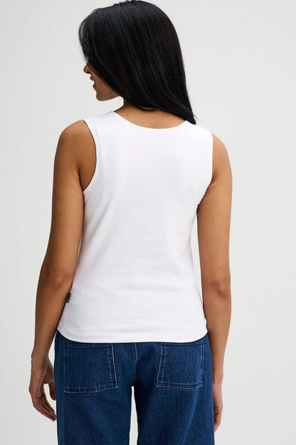 Back view of a woman wearing the Brompton Tank by Message Factory in White, a crew neck tank with wide straps and a slim fit in organic cotton. She is wearing it with jeans and standing in front of a white background. 