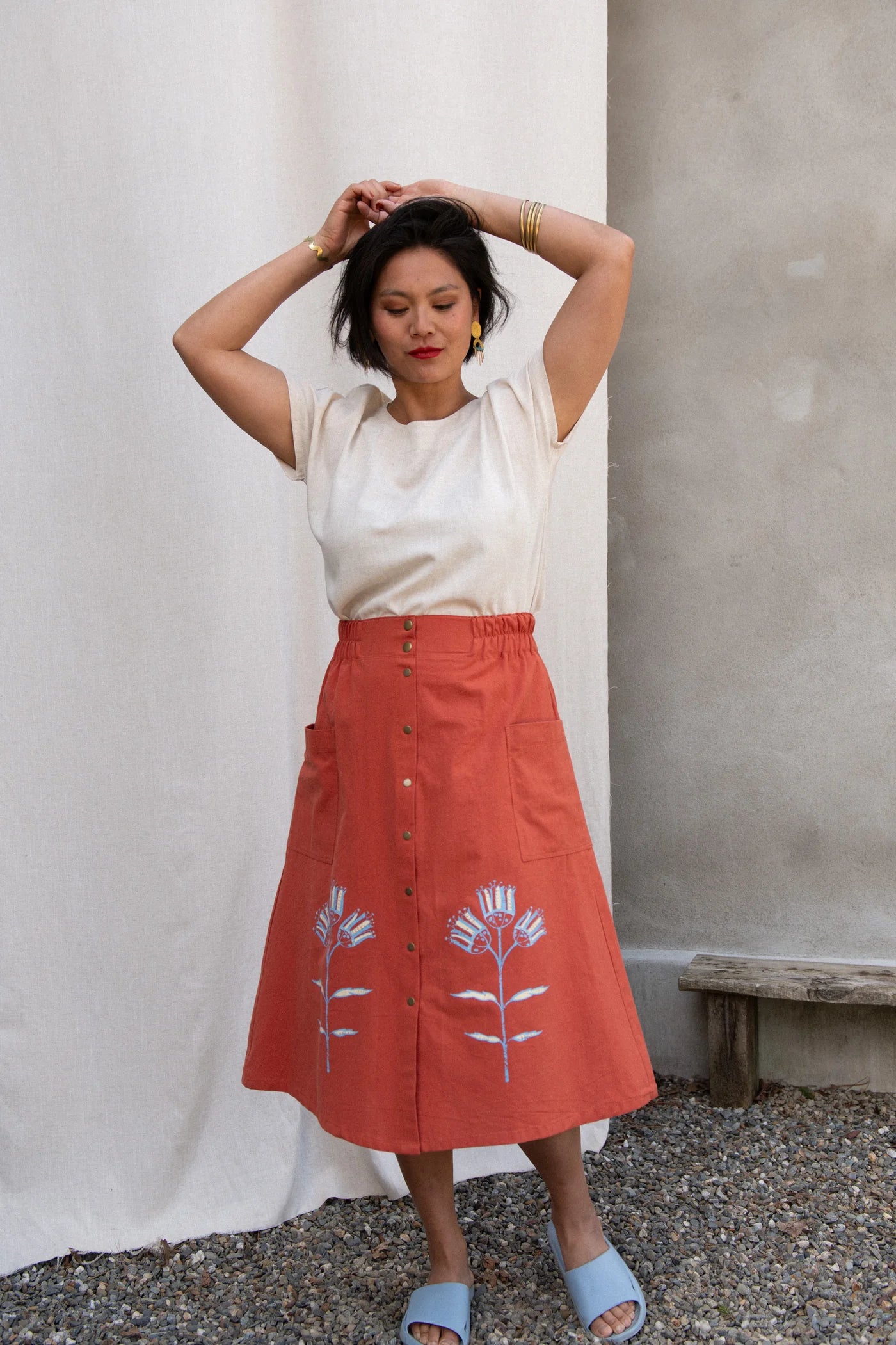 A woman wearing the Nymphea Screenprinted Skirt by Kazak in Red Pysanka with a cream top, she is standing outside in front of white curtain. 