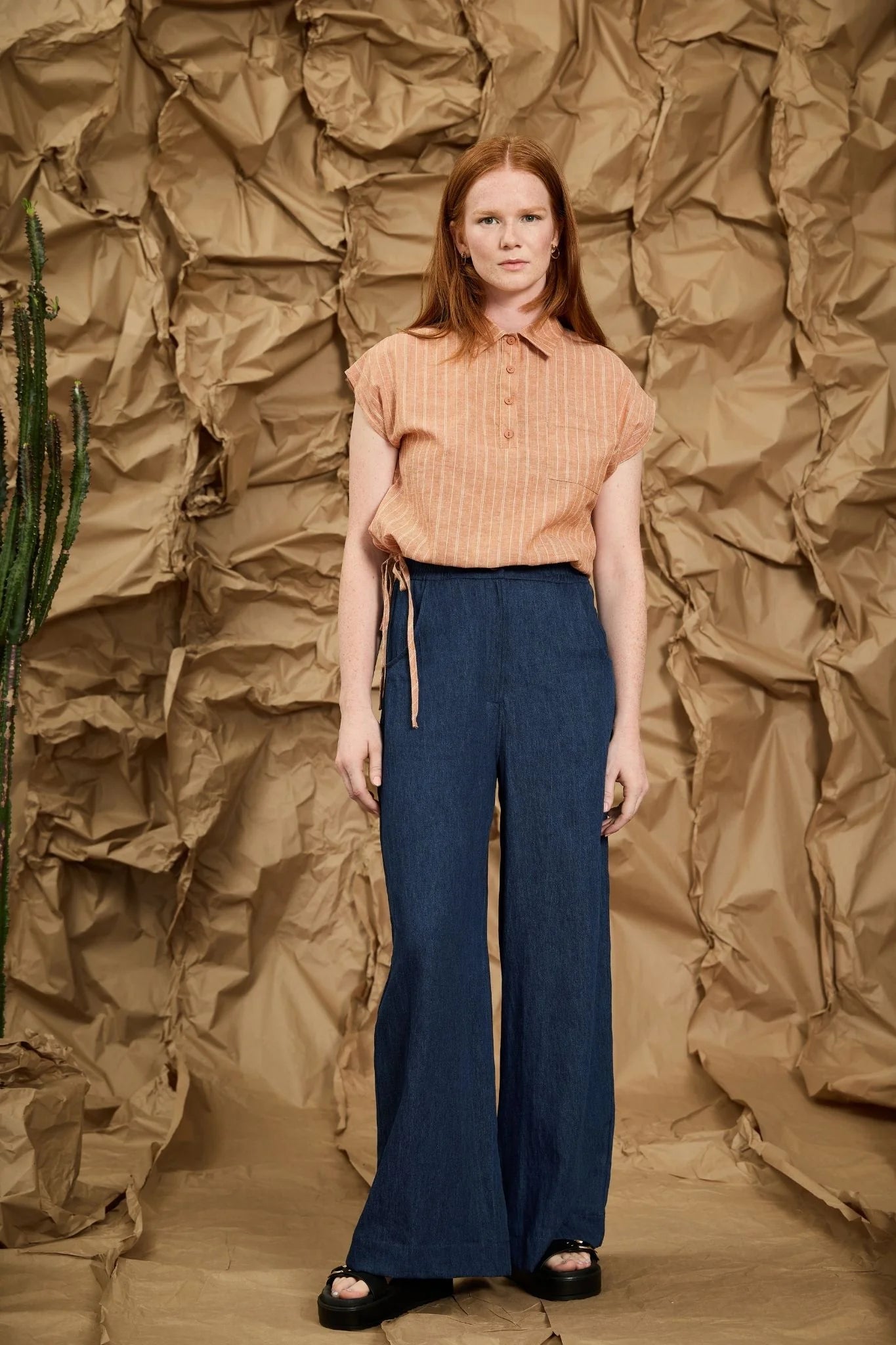 A woman wearing the Matisse Trousers by Melow in Denim, full length wide leg pants with a zipper front and a high waist with elastic at the back and side and back patch pockets. She is wearing them with a peach top and standing in front of a textured brown backdrop