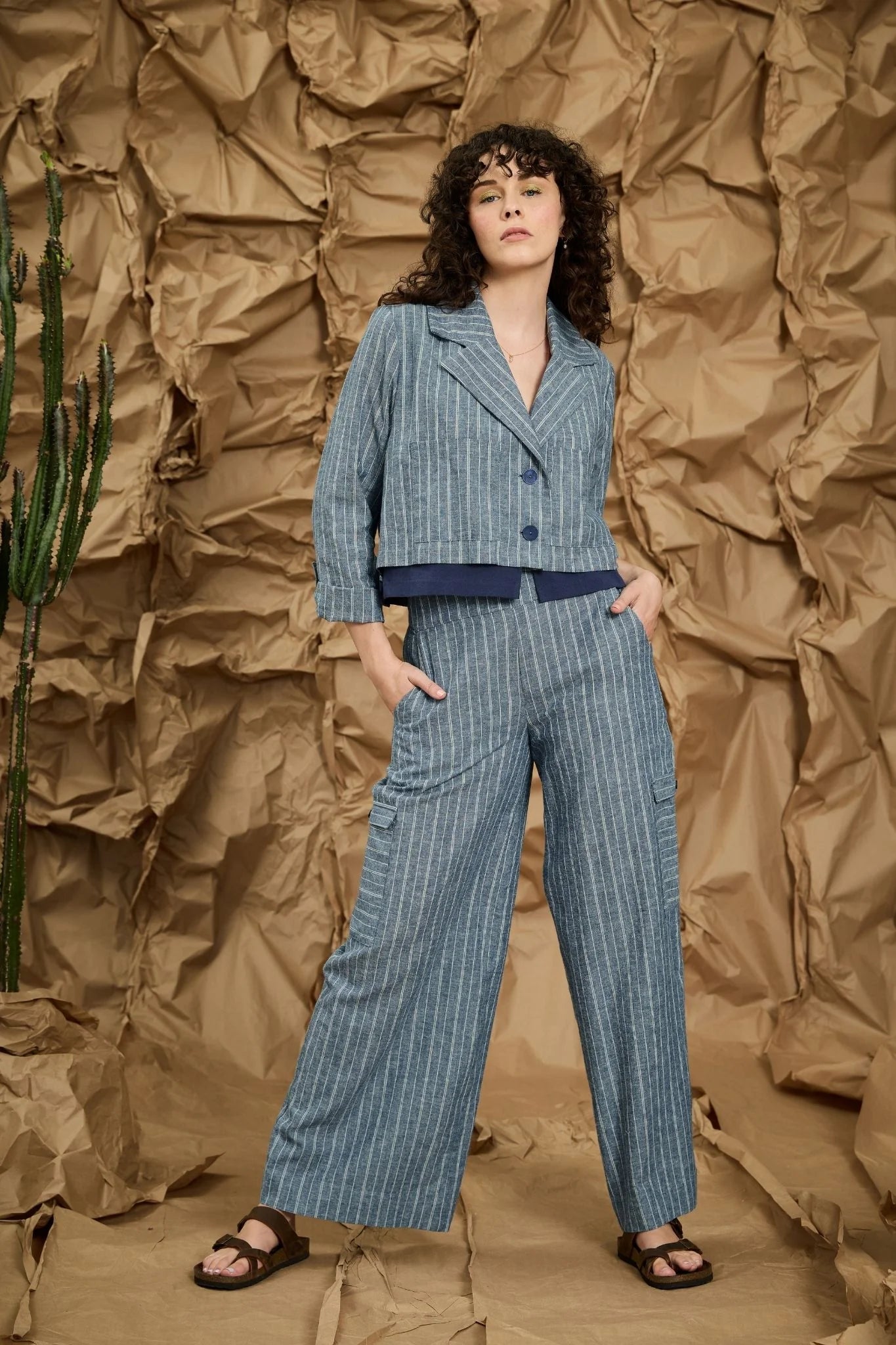 A women wearing the Maroua Jacket by Melow in Blue stripe, a cropped jacket with a tailored collar, two buttons, sleeves with a button cuff detail, and a contrasting lining band. She is wearing the matching Maxime Pants and standing against a textured brown background