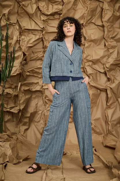 A women wearing the Maroua Jacket by Melow in Blue stripe, a cropped jacket with a tailored collar, two buttons, sleeves with a button cuff detail, and a contrasting lining band. She is wearing the matching Maxime Pants and standing against a textured brown background