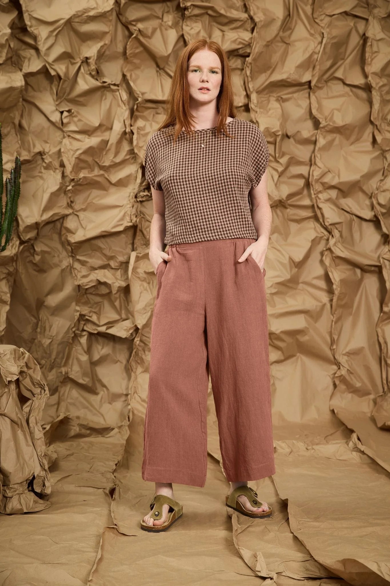 A woman wearing the Marlot Pants by Meow in Rosewood, wide-legged cropped linen pants with a pull on elastic waistband that's smooth at the front. She is wearing them with a patterned top and standing against a textured brown backdrop