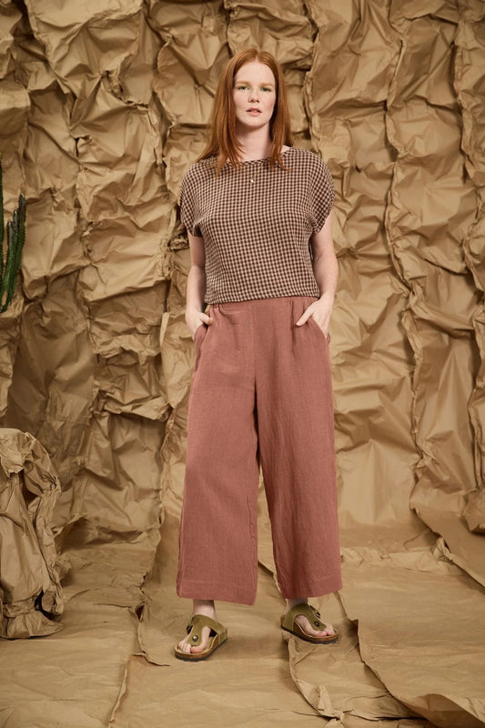 A woman wearing the Marlot Pants by Meow in Rosewood, wide-legged cropped linen pants with a pull on elastic waistband that's smooth at the front. She is wearing them with a patterned top and standing against a textured brown backdrop
