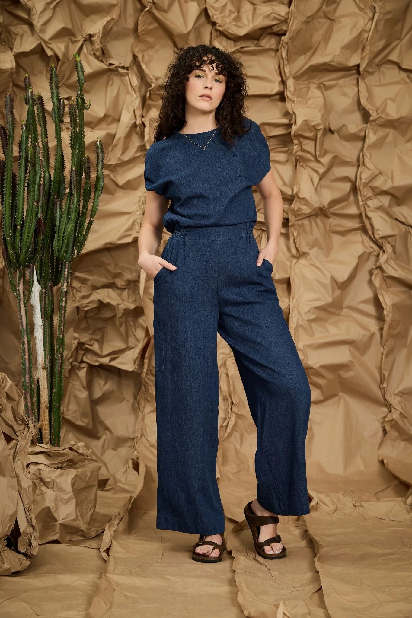 A woman wearing the Michele Jumpsuit by Melow in Denim, with short extended sleeves with gathered cuffs, a defined waist with elastic at the back and sides, an invisible back zip, a chest pocket, and wide full-length legs. She is standing in front of a textured brown background. 