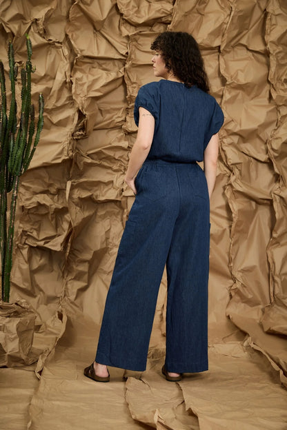 Back view of a woman wearing the Michele Jumpsuit by Melow in Denim, with short extended sleeves with gathered cuffs, a defined waist with elastic at the back and sides, an invisible back zip, a chest pocket, and wide full-length legs. She is standing in front of a textured brown background. 