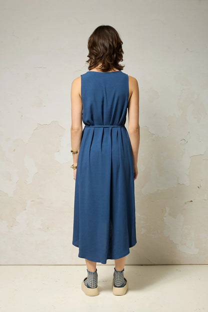 Back view of a woman wearing the Chaos Dress by Cokluch in Denim Blue, a long loose sleeveless dress with a V-neck, matching belt, and hi-low hemline. She is standing against a plain background