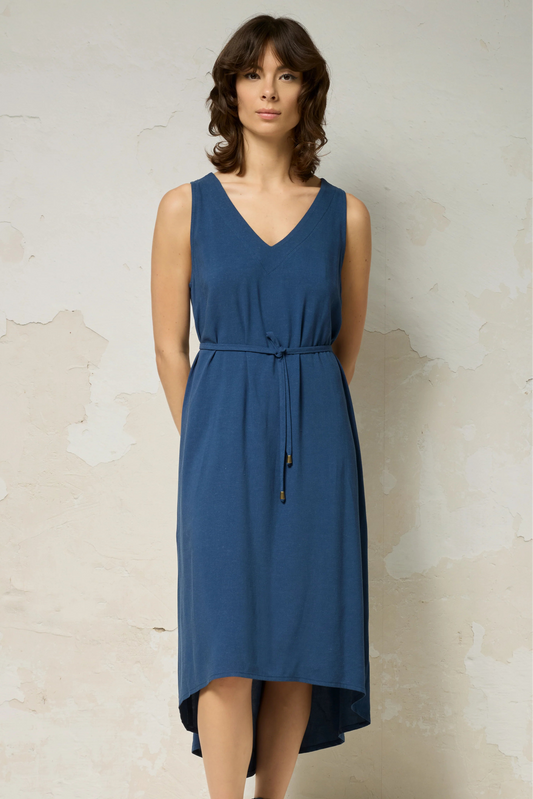 A woman wearing the Chaos Dress by Cokluch in Denim Blue, a long loose sleeveless dress with a V-neck, matching belt, and hi-low hemline. She is standing against a plain background.