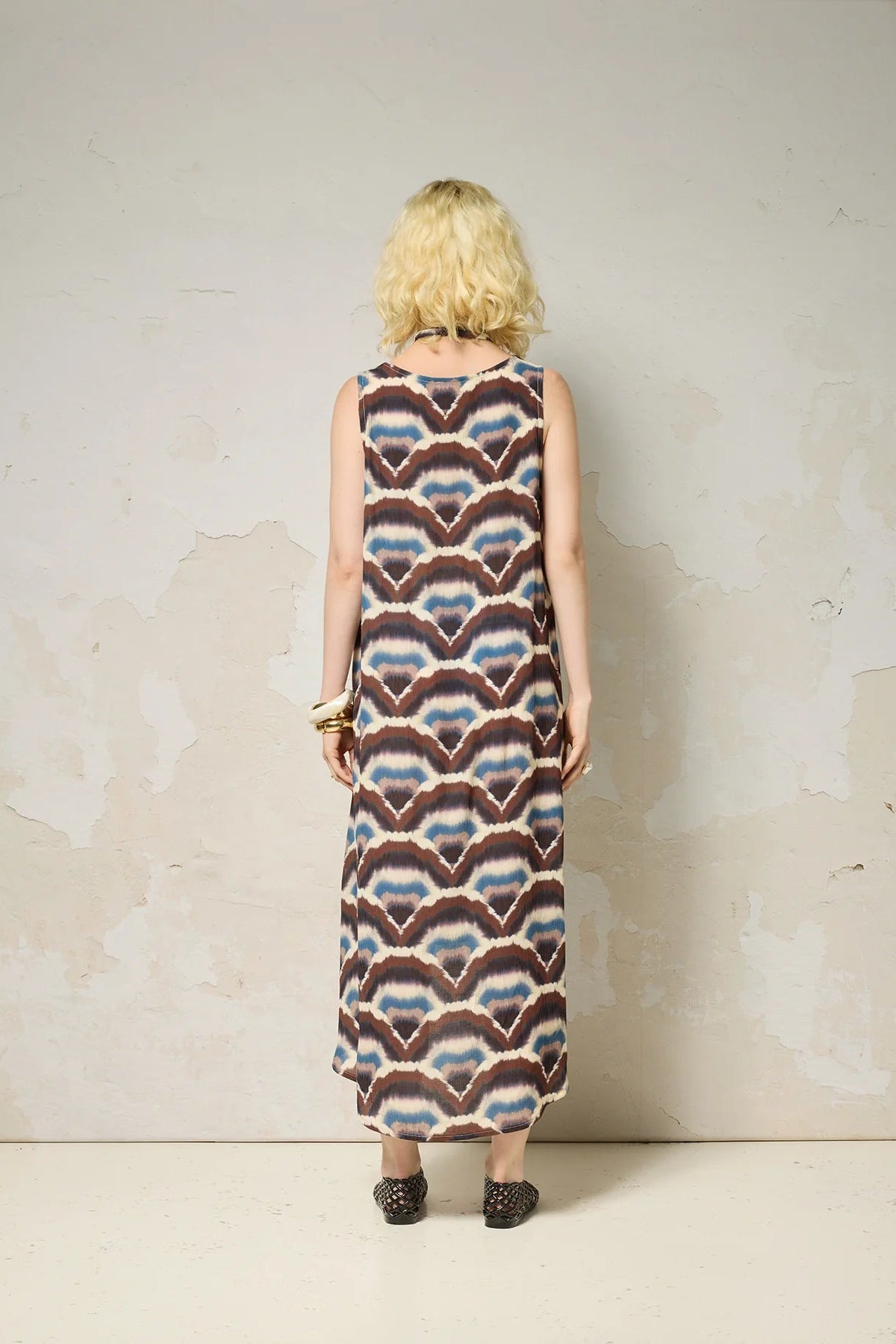 Back view of a woman wearing the Chaos Dress by Cokluch in Feathers print, a long loose sleeveless dress with a V-neck, matching belt, and hi-low hemline. She is standing against a plain background