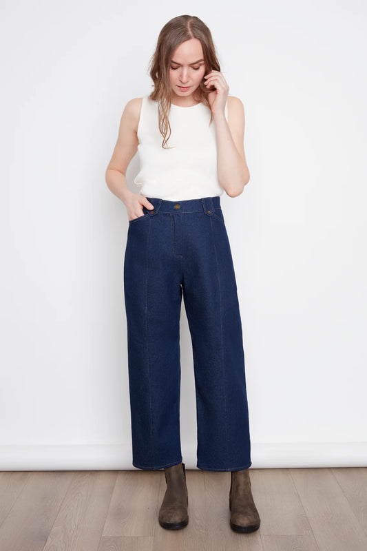 A woman wearing the Alice Straight Leg Pants by Julei in Dark Denim, straight cut jeans with vertical seams down the front and back of the legs, button and zipper closure, and pockets. She is wearing them with a white tank and standing in front of a white background. 