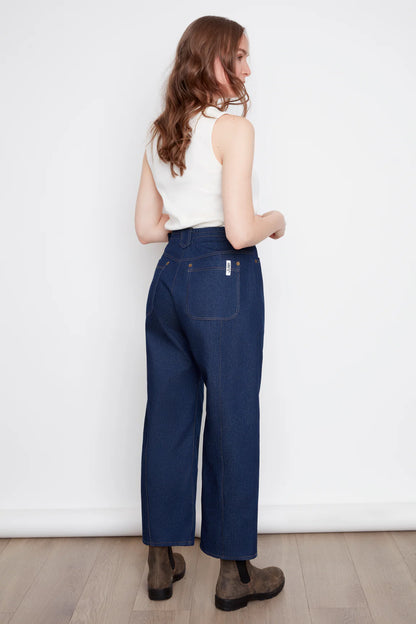 Back view of a woman wearing the Alice Straight Leg Pants by Julei in Dark Denim, straight cut jeans with vertical seams down the front and back of the legs, button and zipper closure, and pockets. She is wearing them with a white tank and standing in front of a white background. 
