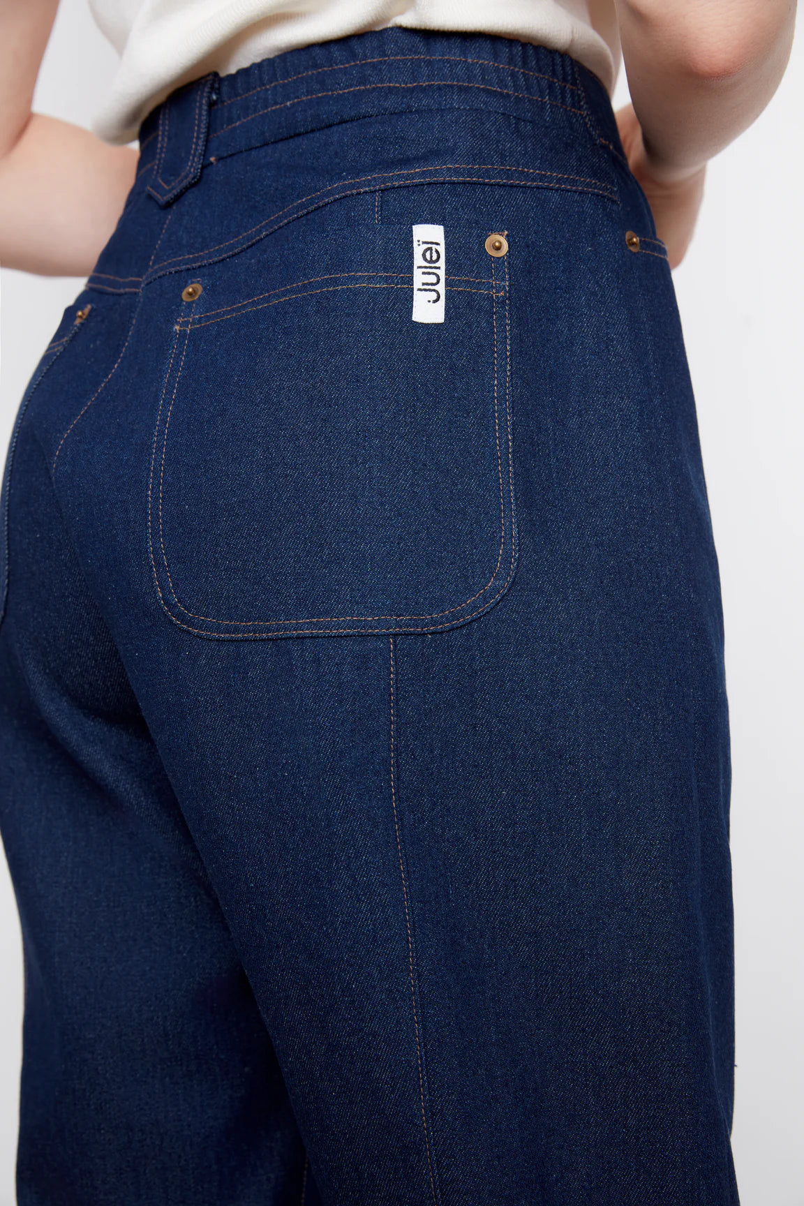Waist down back view of a woman wearing the Alice Straight Leg Pants by Julei in Dark Denim, straight cut jeans with vertical seams down the front and back of the legs, button and zipper closure, and pockets. She is wearing them with a white tank and standing in front of a white background. 