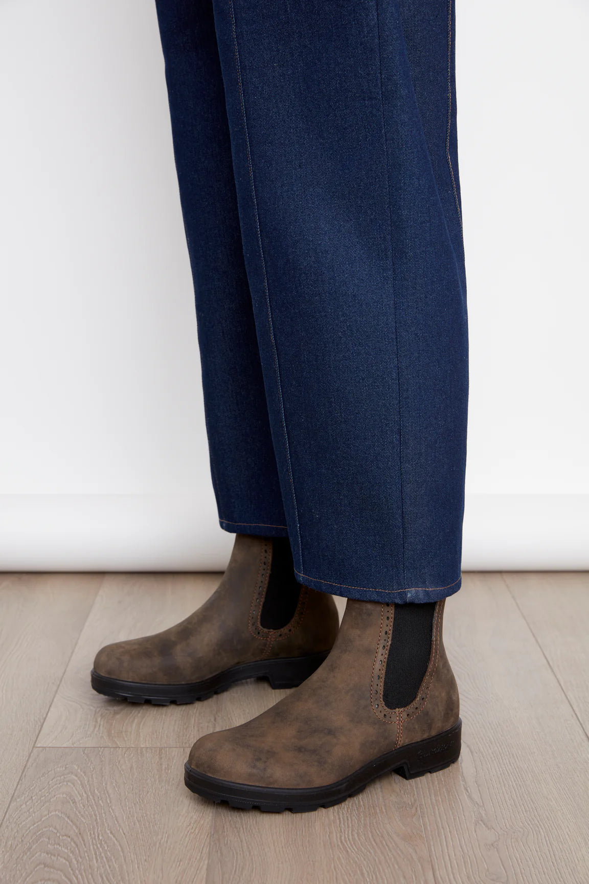 Lower view of a woman wearing the  Alice Straight Leg Pants by Julei in Dark Denim, straight cut jeans with vertical seams down the front and back of the legs, button and zipper closure, and pockets. She is standing in front of a white background. 