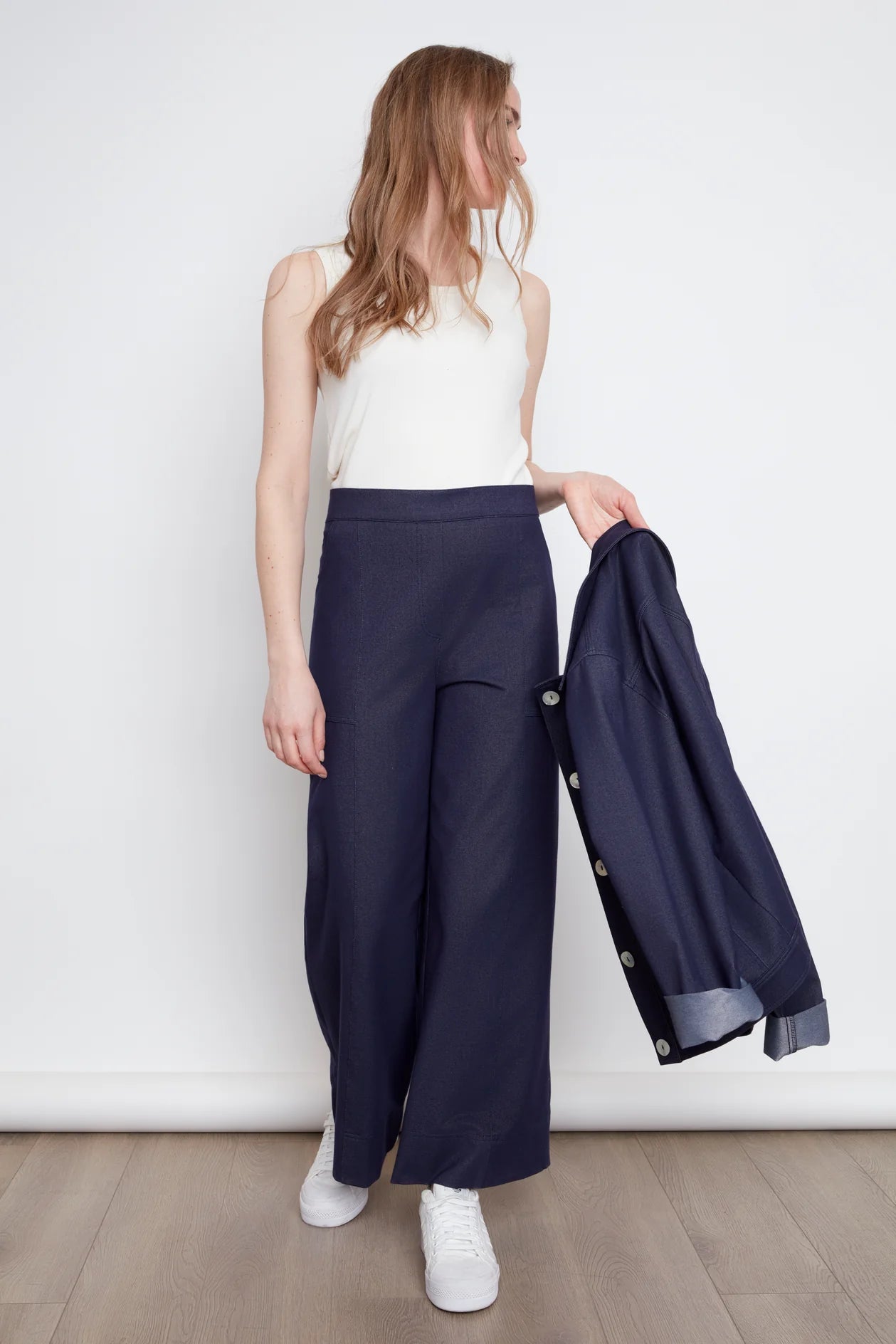 A woman wearing the Lucille Tank by Julei in Natural White, a ribbed tank with a round neck. She is wearing it with denim pants and holding a denim jacket in her hand. 