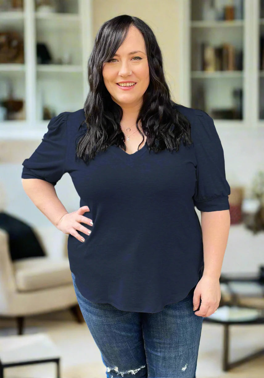 A woman wearing the Darling Tee by Dotty in Navy, featuring a v-neck, pleated shoulders, and elbow length sleeves, with jeans. She is standing indoors in front of a couch.
