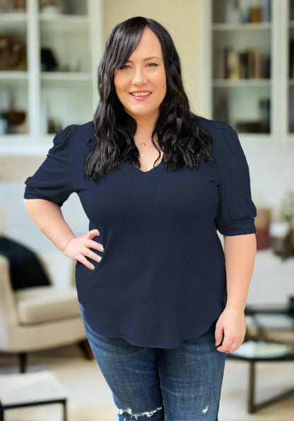 A woman wearing the Darling Tee by Dotty in Navy, featuring a v-neck, pleated shoulders, and elbow length sleeves, with jeans. She is standing indoors in front of a couch.