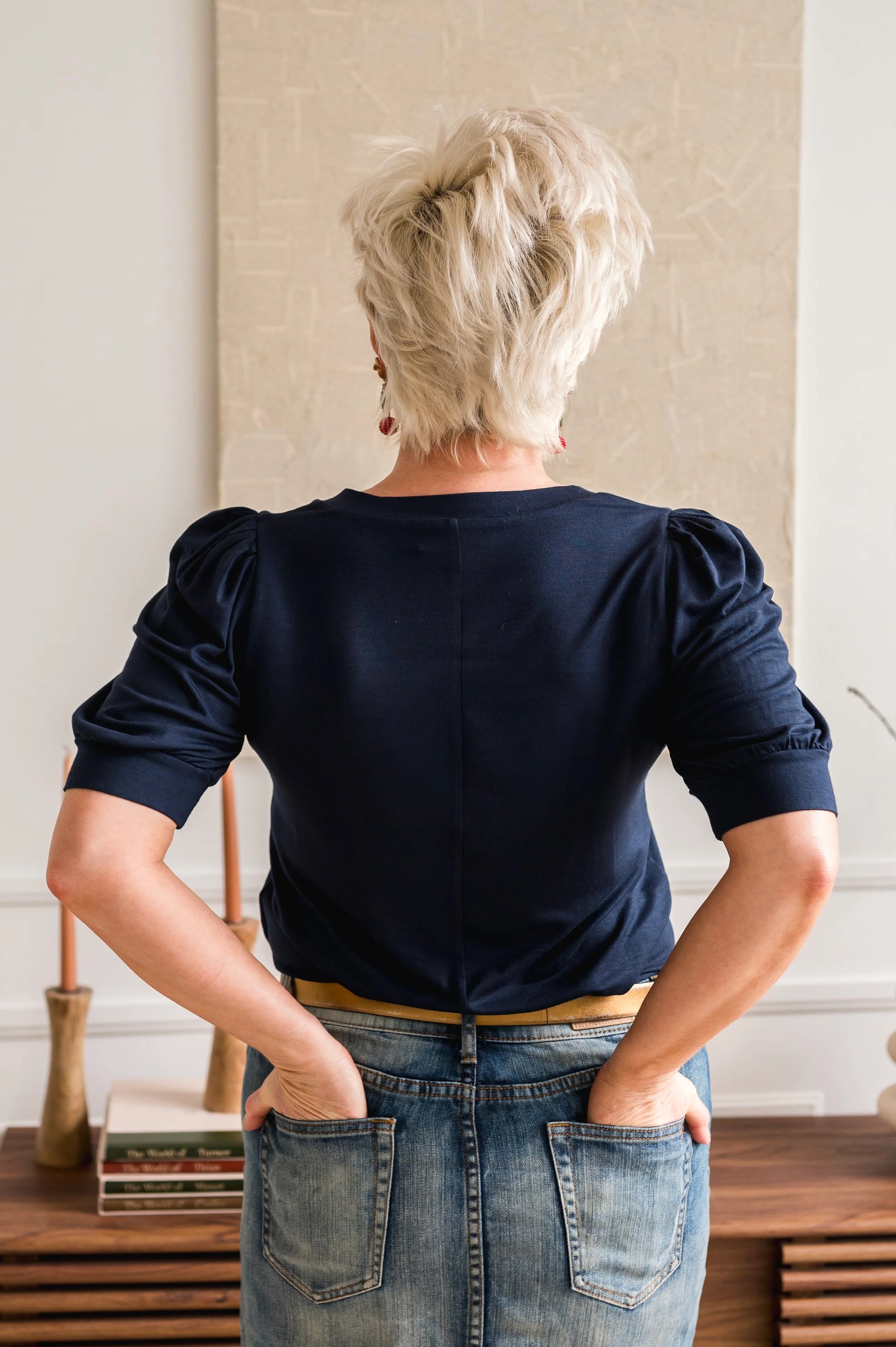 Back view of a woman wearing the  Darling Tee by Dotty in Navy, featuring a v-neck, pleated shoulders, and elbow length sleeves, with a jean skirt. She is standing in front of table. 