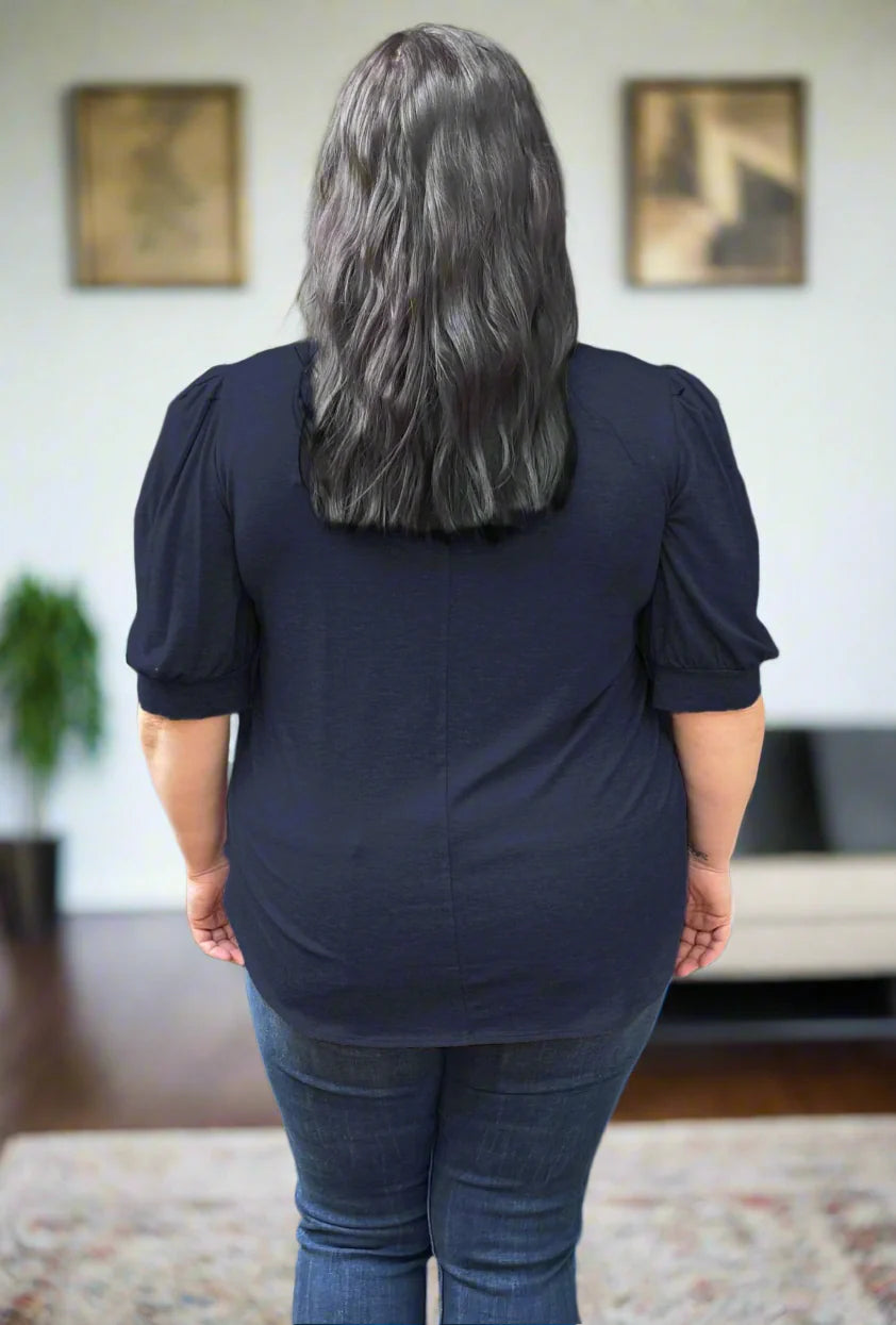 Back view of a woman wearing the  Darling Tee by Dotty in Navy, featuring a v-neck, pleated shoulders, and elbow length sleeves, with a jeans. She is standing in front of a couch.
