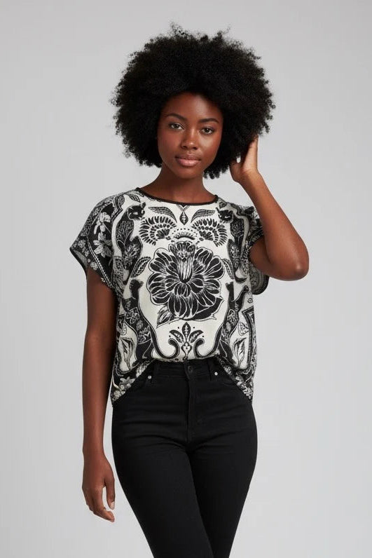A woman wearing the Malo T-shirt from Kollontai, featuring a floral and panther print in black-on-ivory on the front, cap sleeves, and a solid black back. She is wearing it with black pants and standing against a white background. 