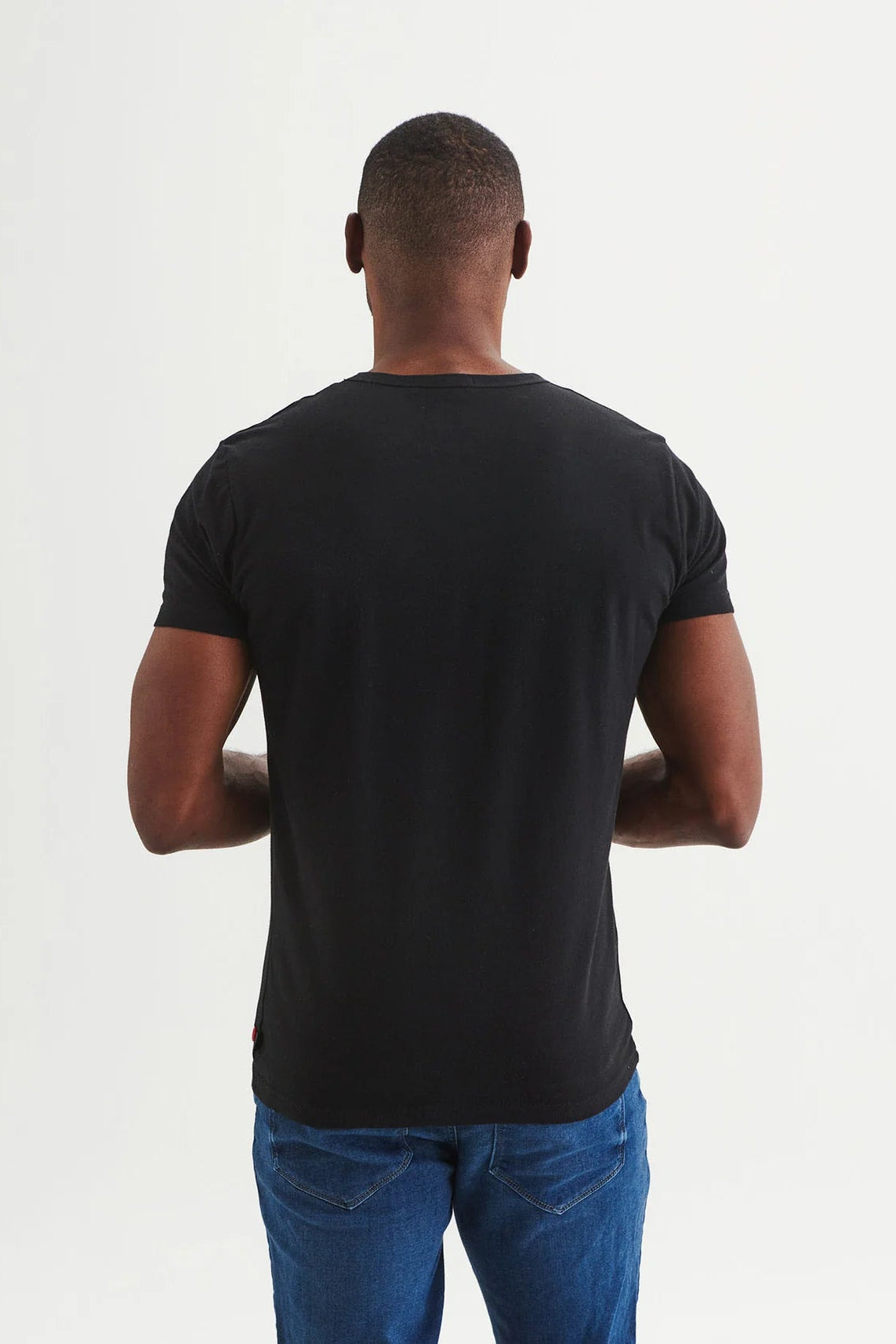 Back view of a man wearing the Sommet T-shirt by Message Factory in Black, a classic t-shirt with cycling-inspired graphic in white. He is wearing it with jeans and standing in front of a white background. 