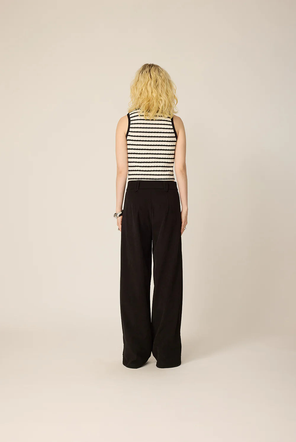 Back view of a woman wearing the Hebe Shirt from Cokluch, a black and white striped sleeveless top with a round neck. She is wearing it with black pants against a beige background