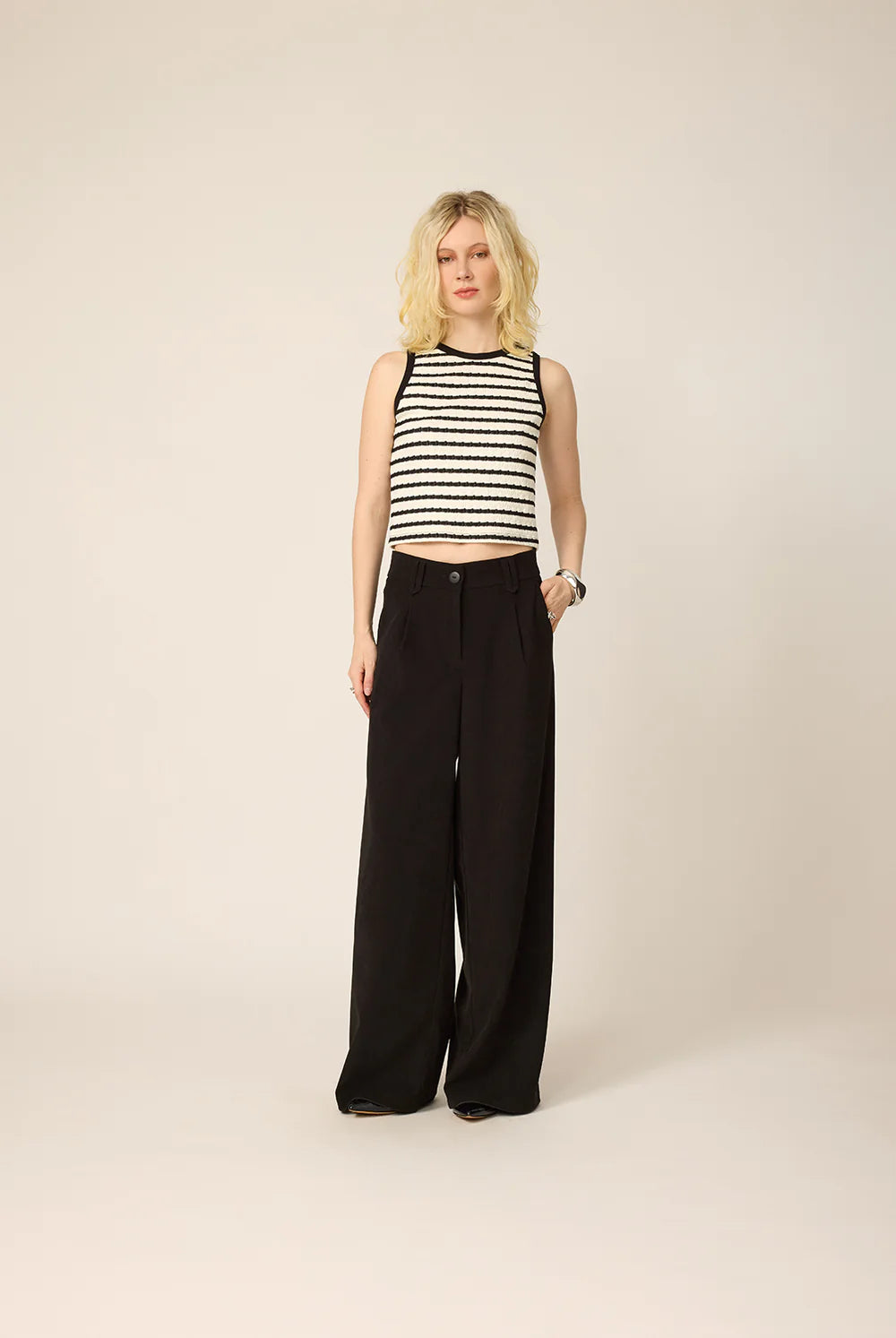 A woman wearing the Hebe Shirt from Cokluch, a black and white striped sleeveless top with a round neck. She is wearing it with black pants against a beige background