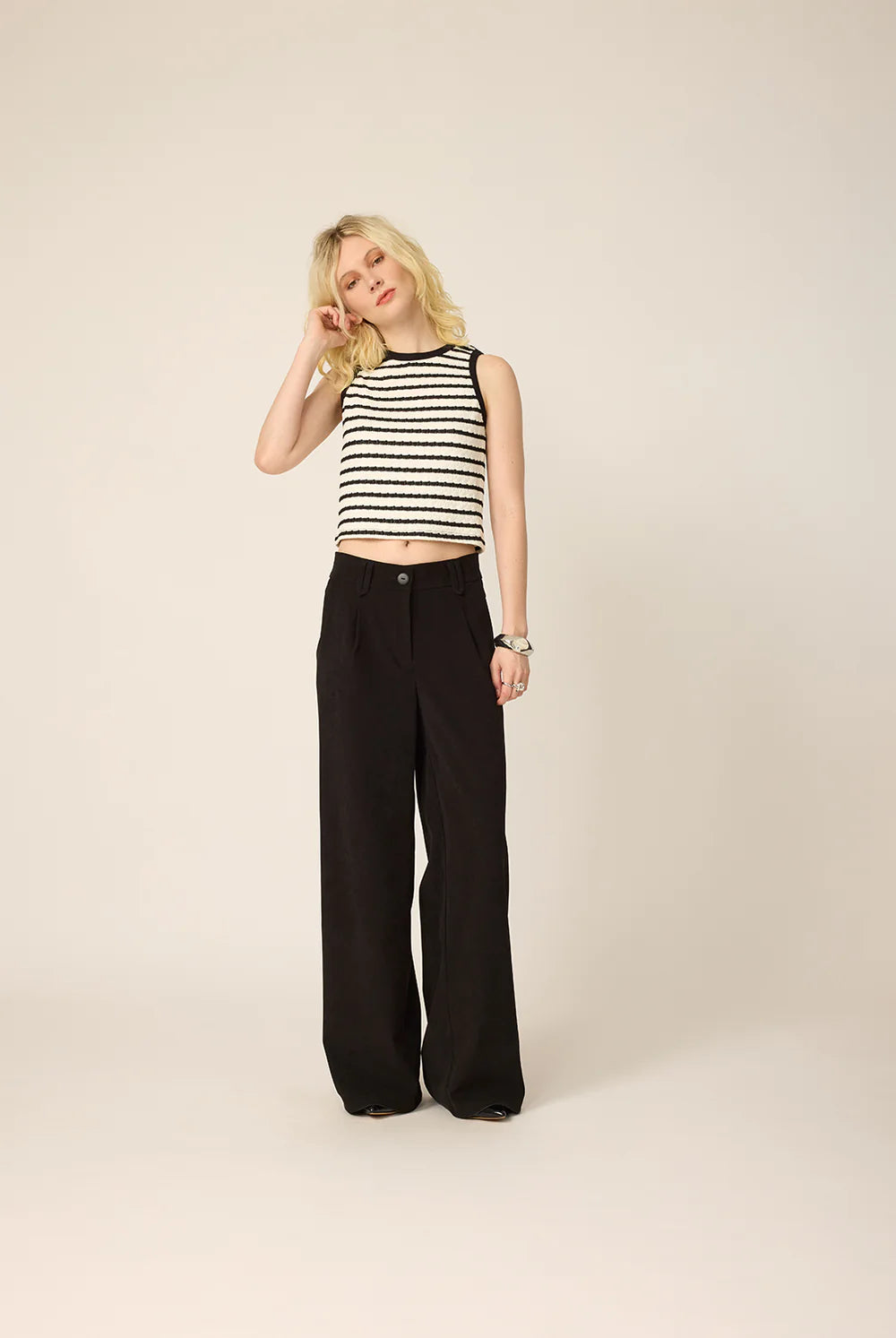 A woman wearing the Hebe Shirt from Cokluch, a black and white striped sleeveless top with a round neck. She is wearing it with black pants against a beige background