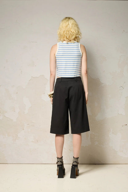 A woman wearing the Loke Shorts from Cokluch in Black, long Bermuda shorts with wide legs, a slightly dropped waist, and pointed belt loops. She is wearing them with a cropped striped tank top and standing in front of a grey wall. 