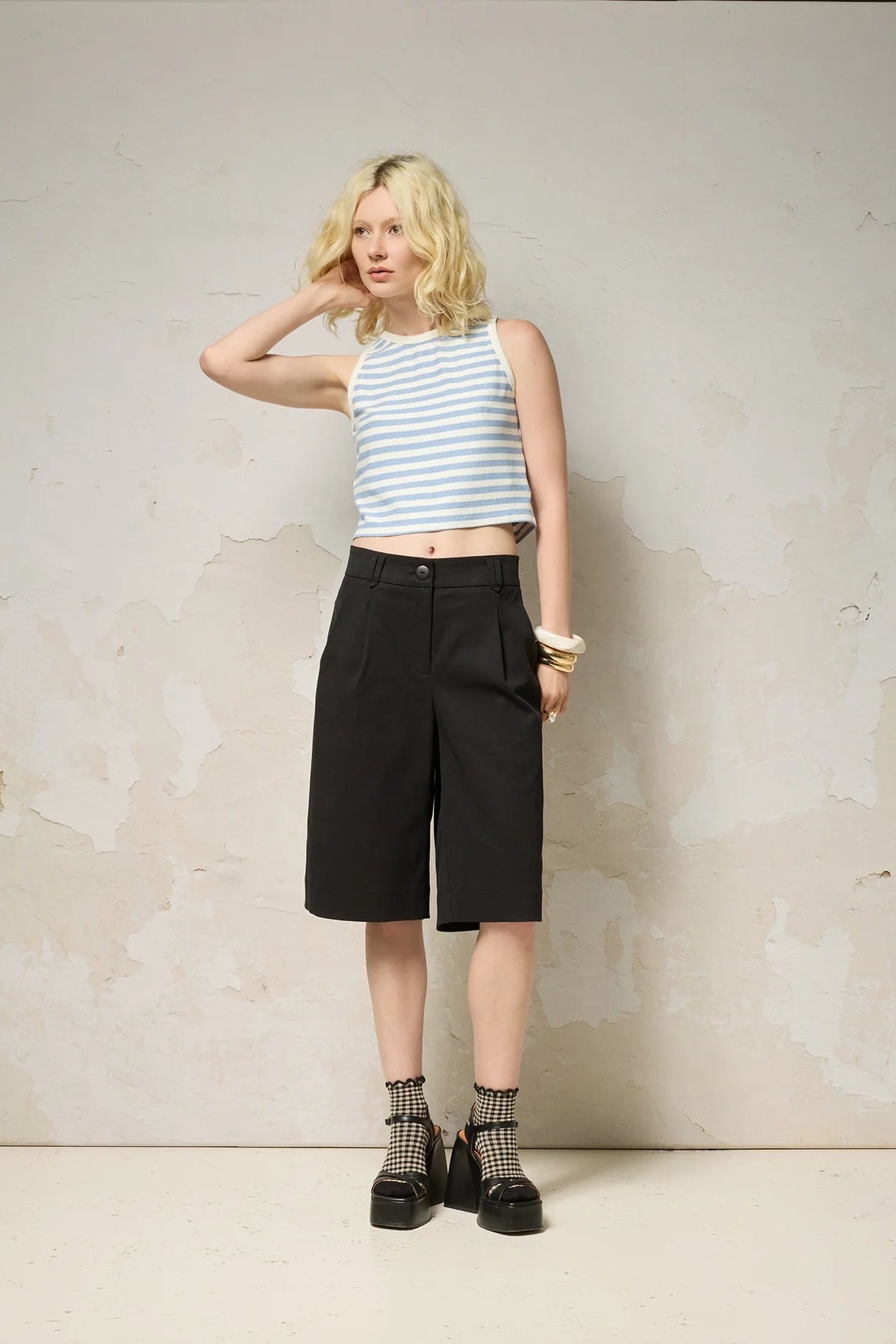 A woman wearing the Loke Shorts from Cokluch in Black, long Bermuda shorts with wide legs, a slightly dropped waist, and pointed belt loops. She is wearing them with a cropped striped tank top and standing in front of a grey wall. 