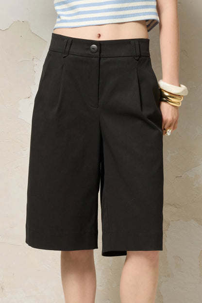 Waist down view of a woman wearing the Loke Shorts from Cokluch in Black, long Bermuda shorts with wide legs, a slightly dropped waist, and pointed belt loops. She is wearing them with a cropped striped top and standing in front of a grey wall. 