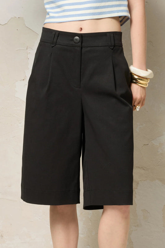 Waist down view of a woman wearing the Loke Shorts from Cokluch in Black, long Bermuda shorts with wide legs, a slightly dropped waist, and pointed belt loops. She is wearing them with a cropped striped top and standing in front of a grey wall. 