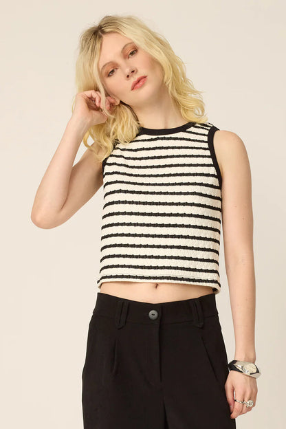 3/4 view of a woman wearing the Hebe Shirt from Cokluch, a black and white striped sleeveless top with a round neck. She is wearing it with black pants against a beige background