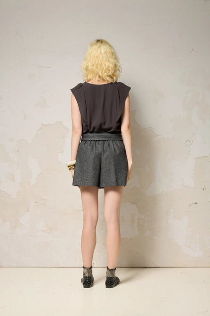 Back view of a woman wearing the Hima Top from Cokluch in Iron, a sleeveless top with shoulder ruffles, a round neck, and hip-length cut. She is wearing it with denim shorts and standing against a textured background. 