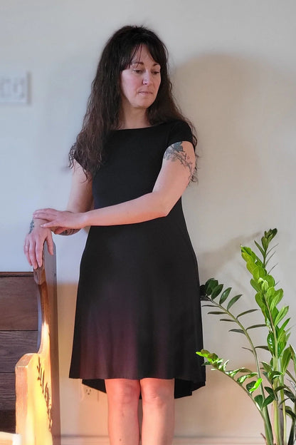 A woman wearing the Hyda dress by Moovment in Charcoal, a short sleeved A-line dress that's reversible back to front. It has a boat neck on one side, a round neck on the other, and hits above the knee. She is standing in a room with a plant and wooden chair.
