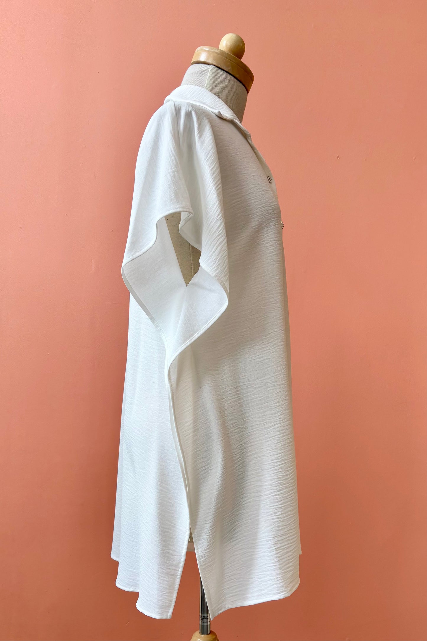 The Geneva Top by Pure Essence in White, a long and loose button front shirt with short sleeves, a classic collar, and high side slits is shown on a mannequin against a coral background. 