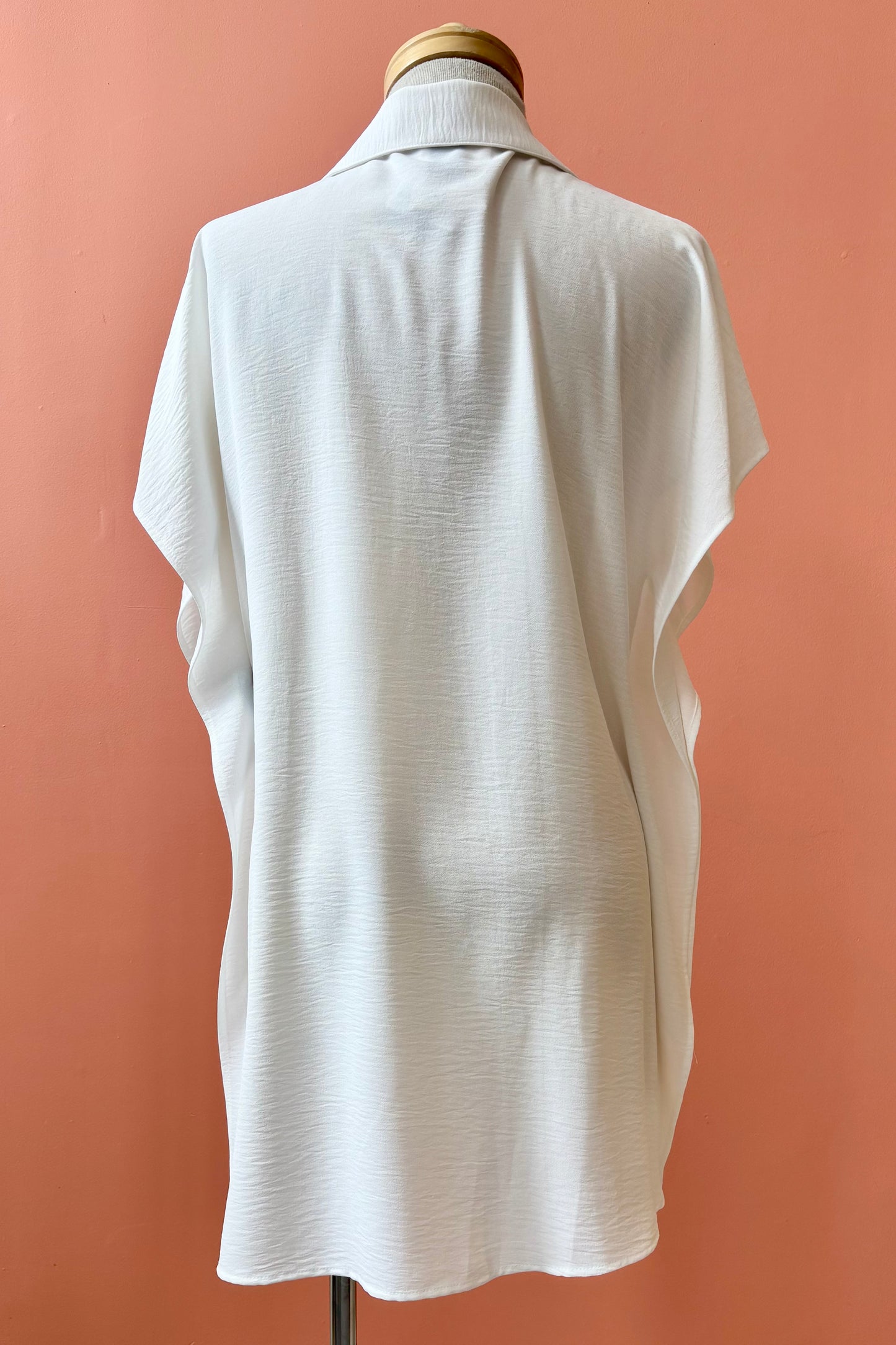 A back view of the Geneva Top by Pure Essence in White, a long and loose button front shirt with short sleeves, a classic collar, and high side slits is shown on a mannequin against a coral background. 
