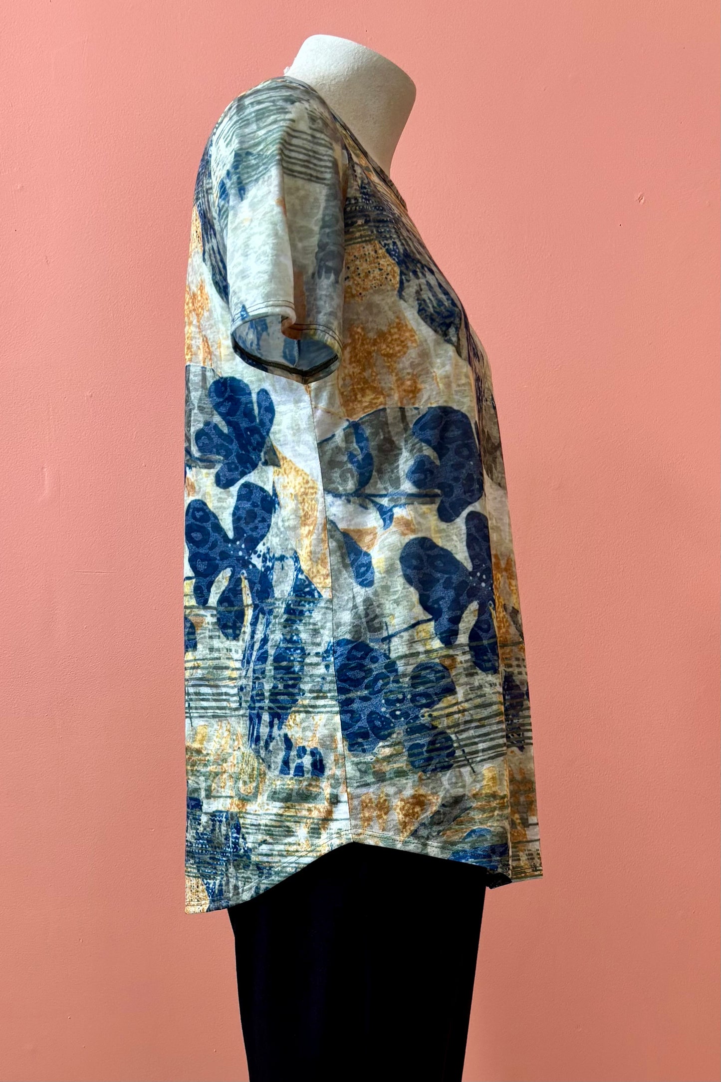 A side view of the Gladys T-shirt by Pure Essence in Golden Multi print is shown on a mannequin against a coral background. 