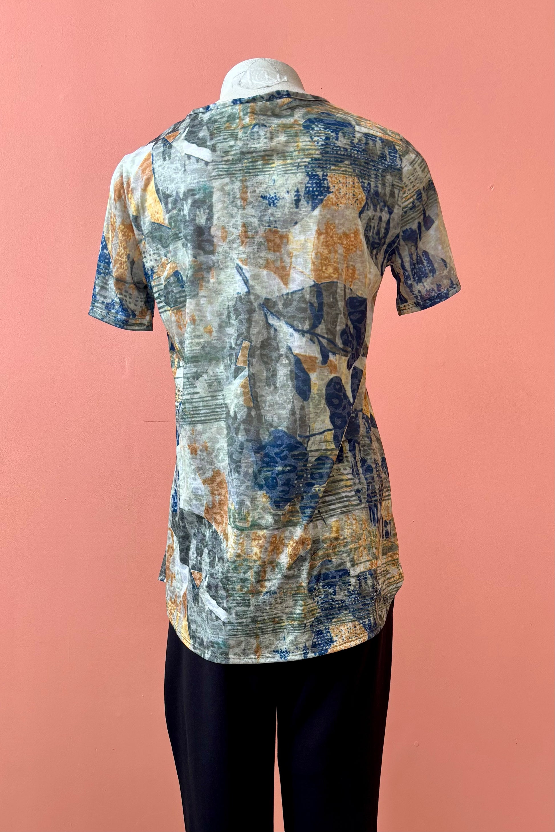 A back view of the Gladys T-shirt by Pure Essence in Golden Multi print is shown on a mannequin against a coral background. 