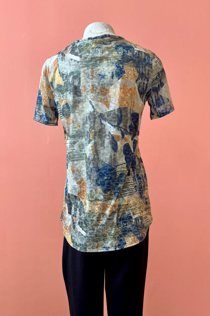A back view of the Gladys T-shirt by Pure Essence in Golden Multi print is shown on a mannequin against a coral background. 