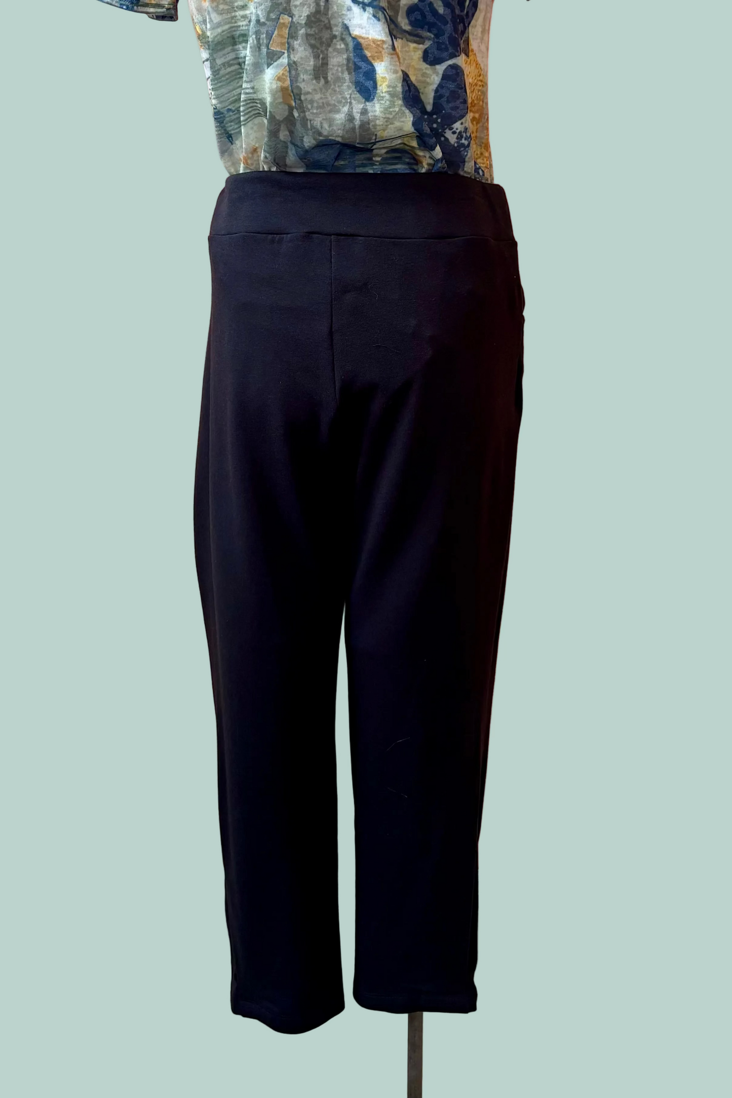 A back view of the Giselle Joggers by Pure Essence in Black are shown on a mannequin with a patterned shirt in front of a turquoise background. 