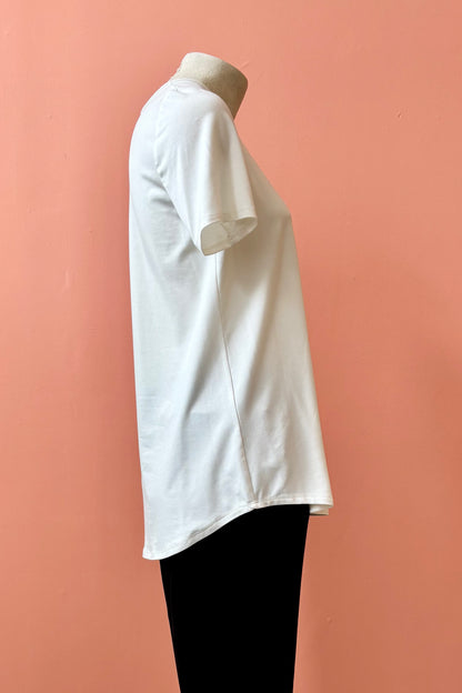 A side view of the Gladys T-shirt by Pure Essence in White is shown on a mannequin against a coral background. 