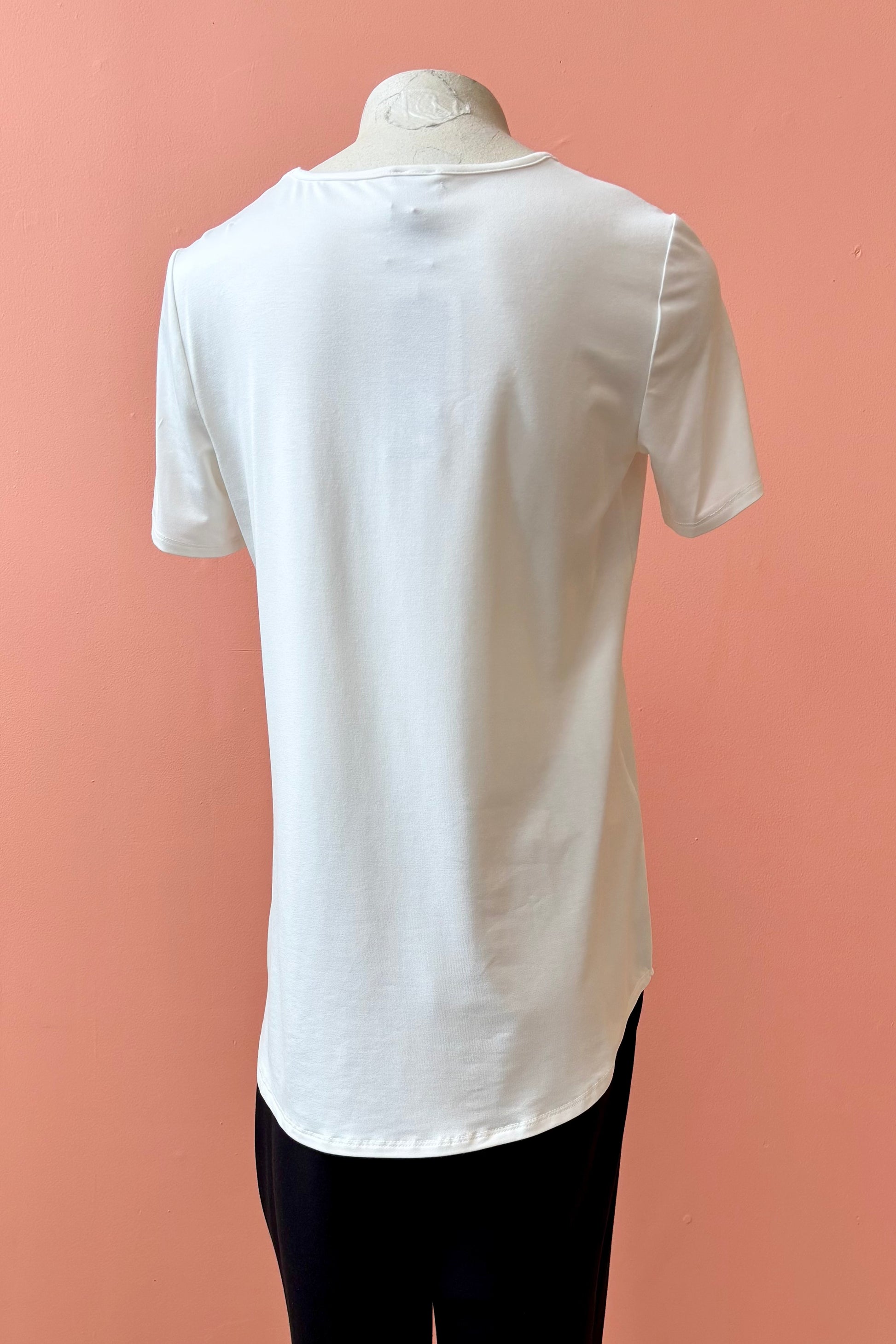 A back view of the Gladys T-shirt by Pure Essence in White is shown on a mannequin against a coral background. 