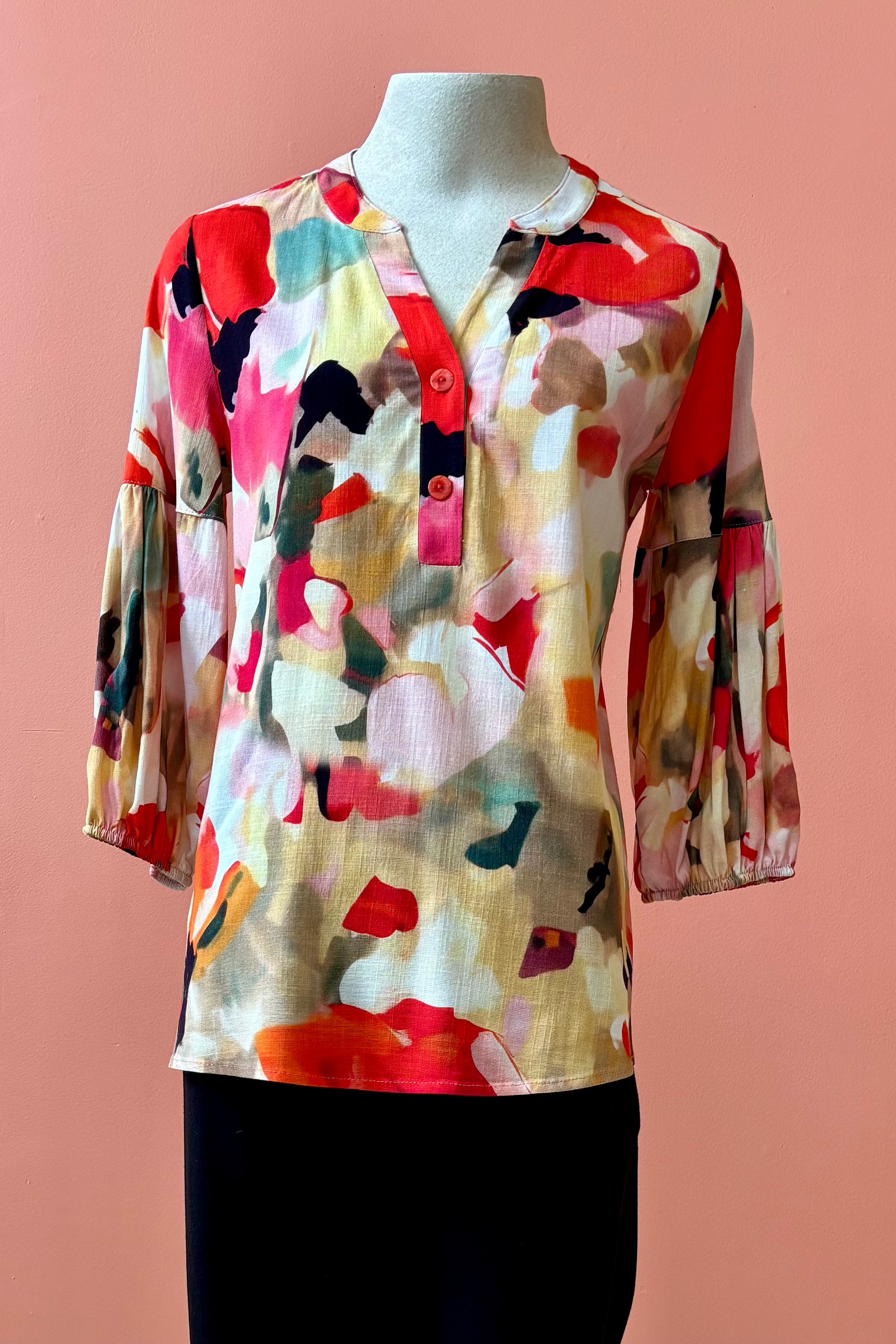 The Grace Blouse by Pure Essence in Coral Meadow, with a v-placket neck with buttons and 7/8 gathered sleeves, is shown with black pants on a mannequin in front of a coral wall. 