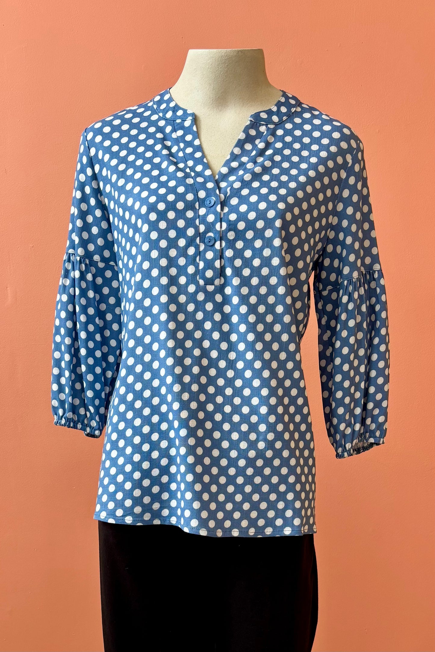 The Grace Blouse by Pure Essence in Chambray Dot, with a v-placket neck with buttons and 7/8 gathered sleeves, is shown with black pants on a mannequin in front of a coral wall. 