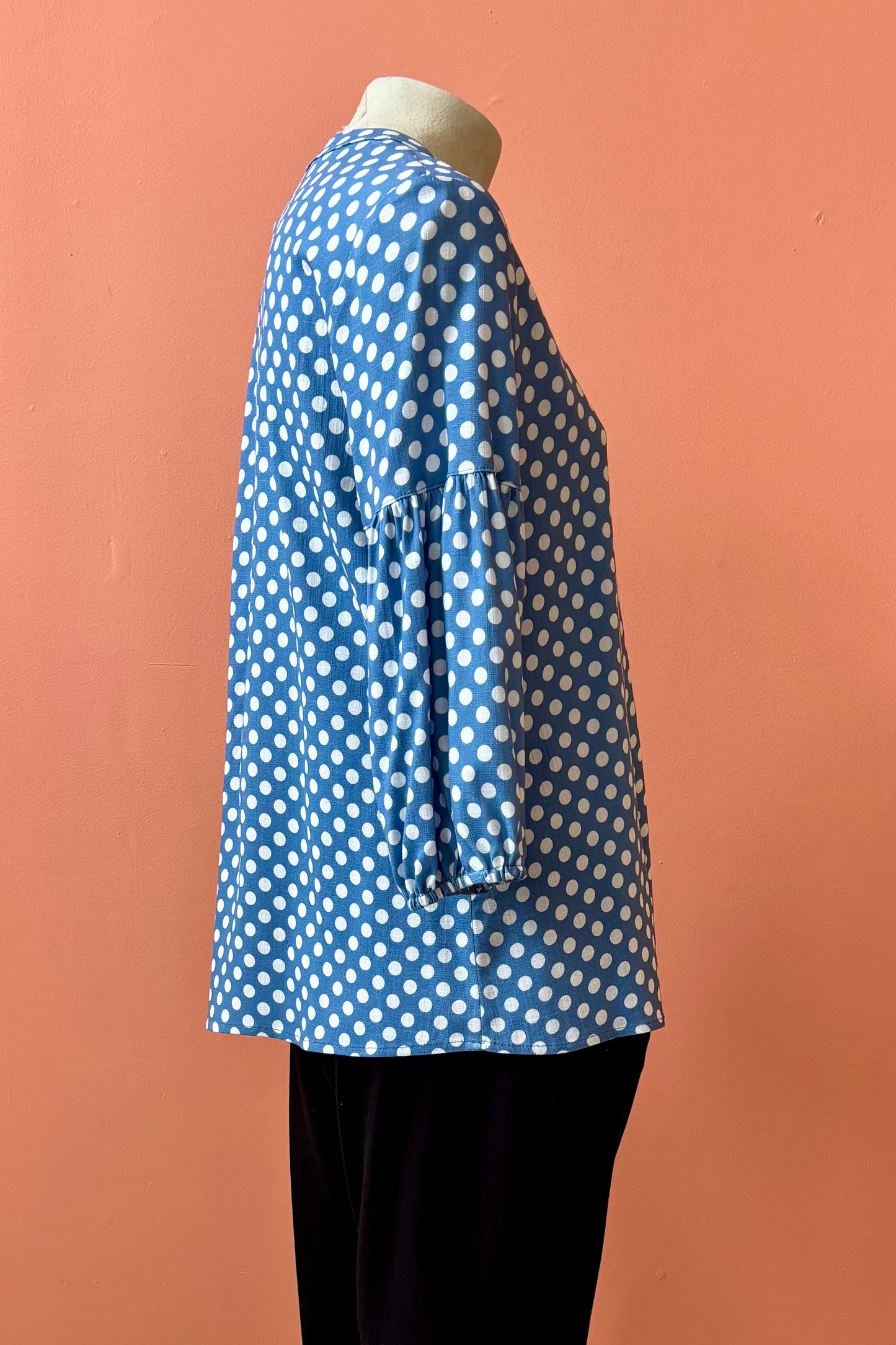 A side view of the Grace Blouse by Pure Essence in Chambray Dot, with a v-placket neck with buttons and 7/8 gathered sleeves, is shown with black pants on a mannequin in front of a coral wall. 