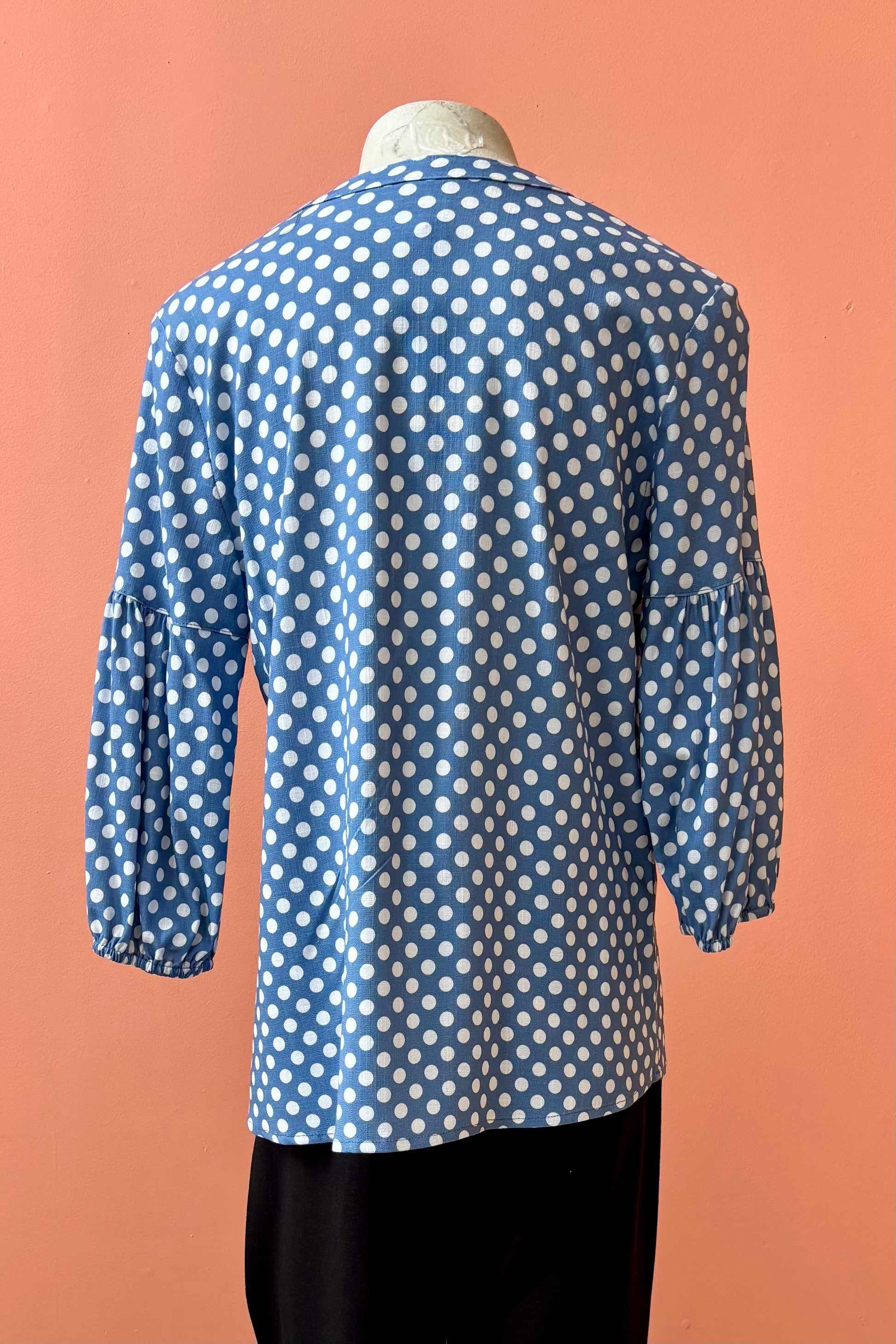 A back view of the Grace Blouse by Pure Essence in Chambray Dot, with a v-placket neck with buttons and 7/8 gathered sleeves, is shown with black pants on a mannequin in front of a coral wall. 