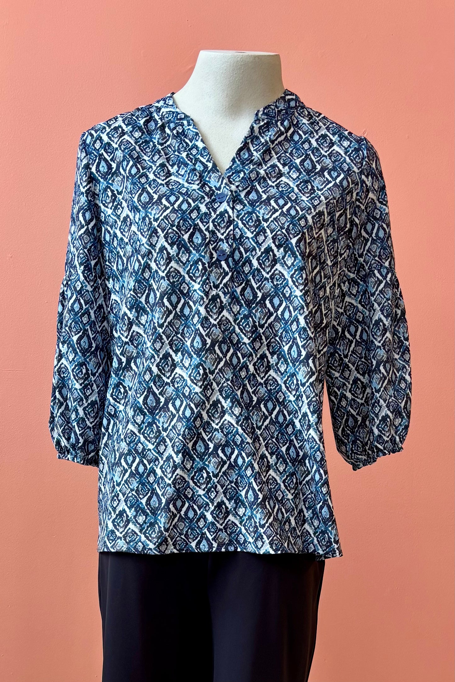 The Grace Blouse by Pure Essence in Blue Lattice, with a v-placket neck with buttons and 7/8 gathered sleeves, is shown with black pants on a mannequin in front of a coral wall. 