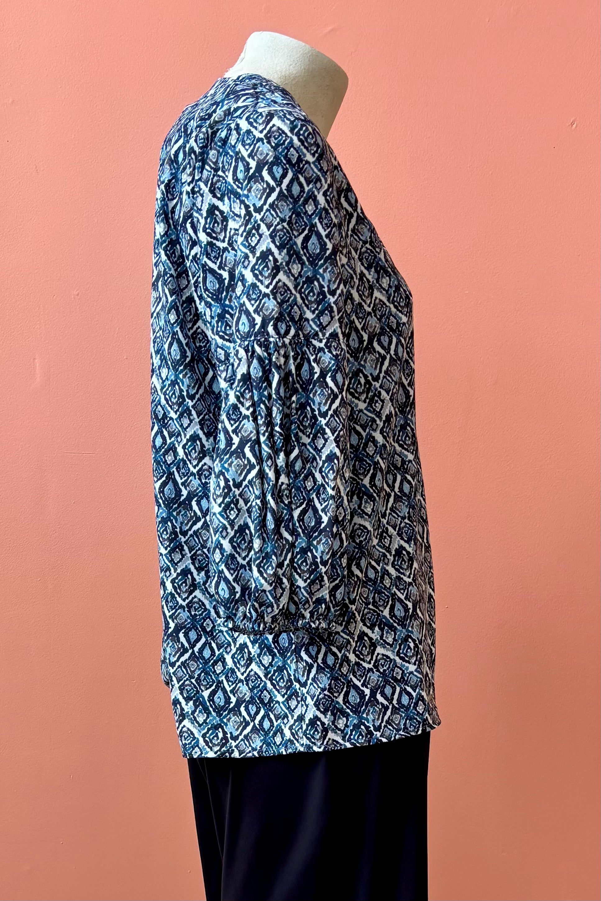A side view of the Grace Blouse by Pure Essence in Blue Lattice, with a v-placket neck with buttons and 7/8 gathered sleeves, is shown with black pants on a mannequin in front of a coral wall. 